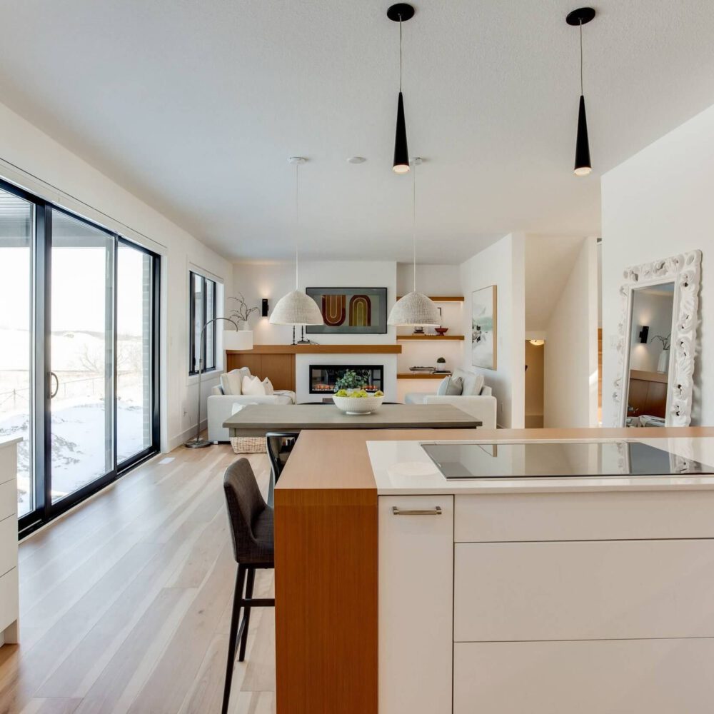 Modern kitchen with white cabinetry, a central island, and pendant lighting, featuring an open layout to a living area with large windows.