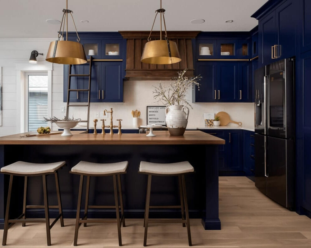 Modern kitchen with navy blue cabinetry, wooden accents, and a central island with three stools. Brass light fixtures add elegance.