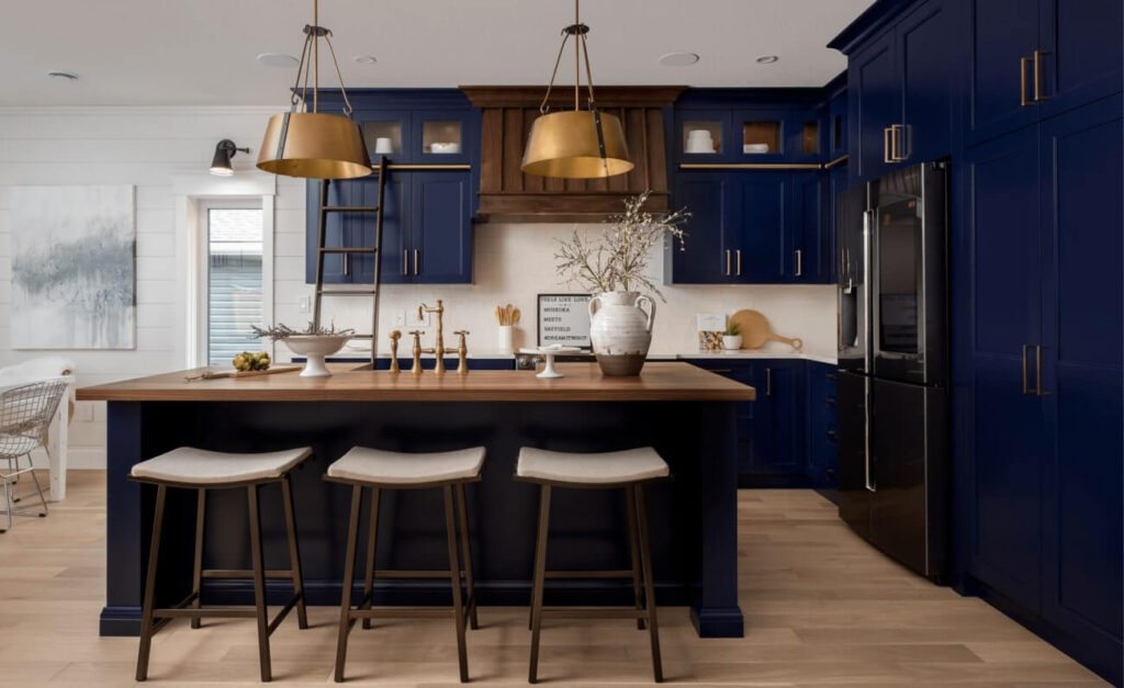 Project_Kitchens_Bayfield2 Modern kitchen with navy blue cabinetry, wooden accents, and a central island with three stools. Brass light fixtures add elegance.