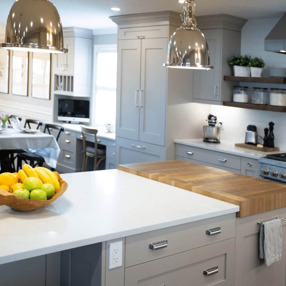 Modern kitchen with a wooden bowl of fruits on a countertop, stainless steel appliances, and stylish pendant lighting.