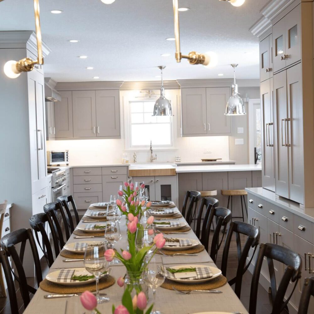 Modern kitchen with a long dining table set with plates and glasses, featuring light gray cabinetry and floral centerpieces.