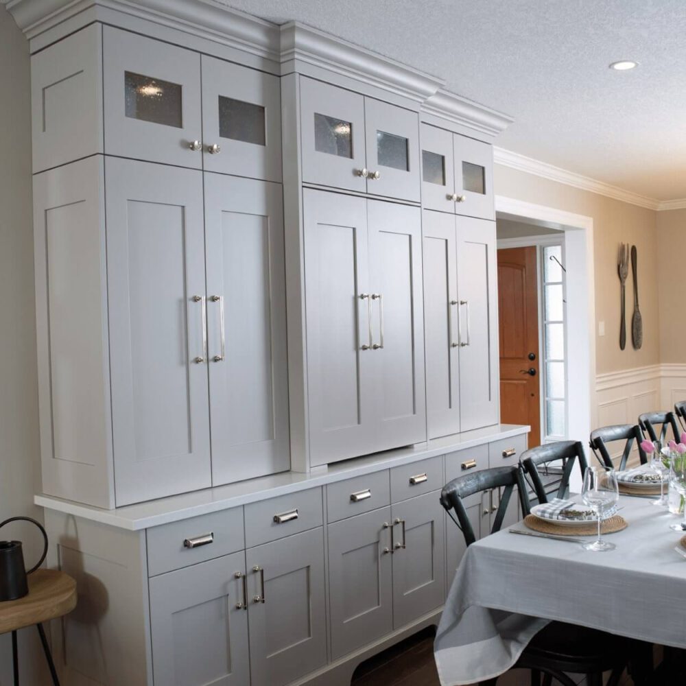 Gray kitchen cabinetry with glass panel accents and a long dining table set with flowers and tableware in a well-lit room.