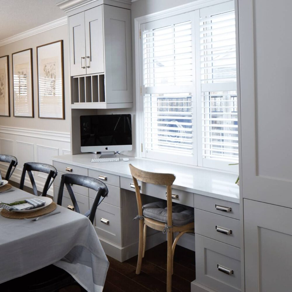 Modern kitchen interior featuring a long dining table, gray cabinetry, and a built-in workspace with a computer and window shutters.