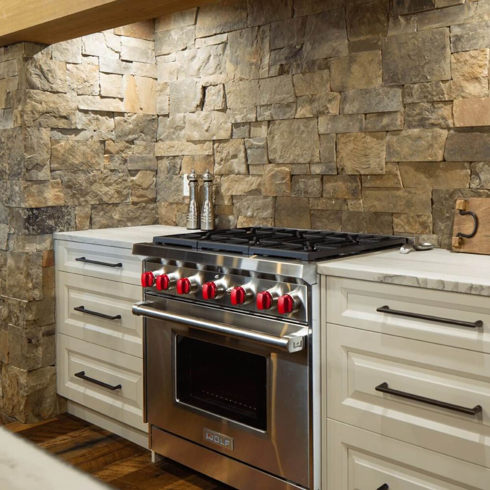 Stainless steel gas range with red knobs installed in a kitchen with stone wall and white cabinetry.