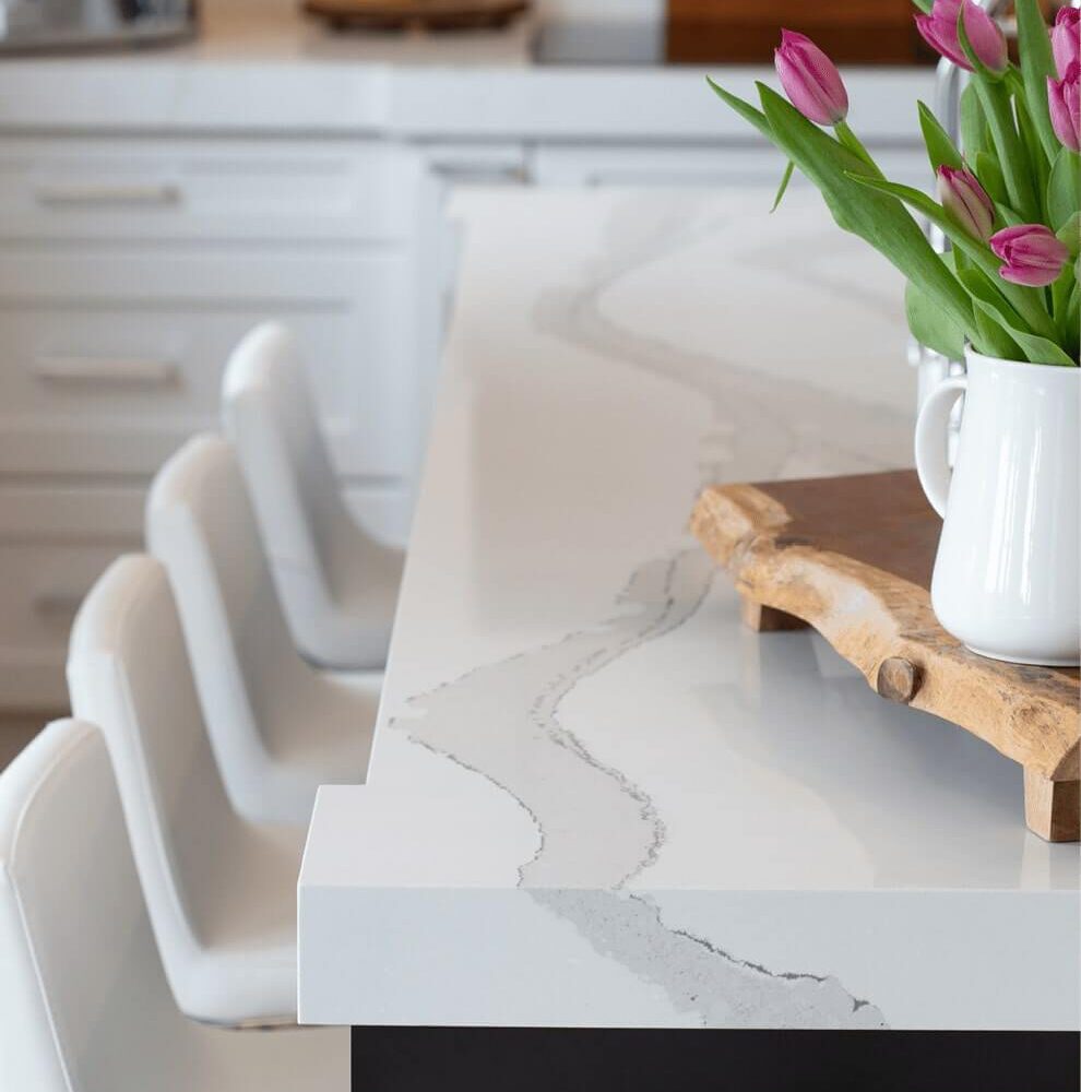 White kitchen countertop with a marble pattern, featuring a wooden cutting board and a vase of tulips. Three white bar stools are positioned nearby.