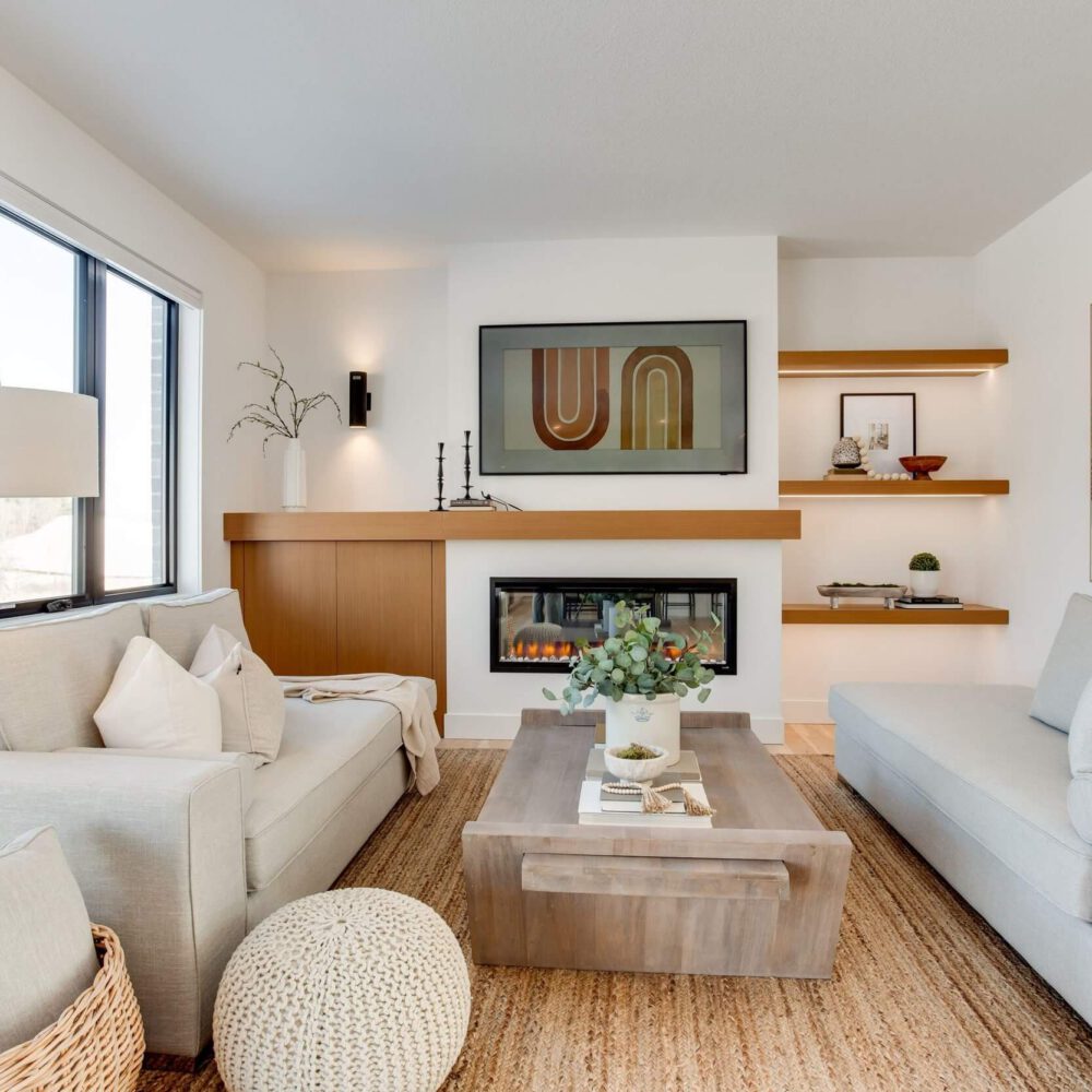 Modern living room with two light gray sofas, a wooden coffee table, and a wall-mounted fireplace, featuring decorative shelves and artwork.