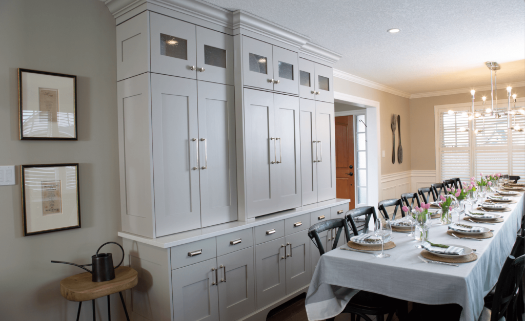 Project_Kitchens_Eastbridge11 Gray kitchen cabinets with glass panels and a long dining table set with flowers and tableware in a well-lit dining area.