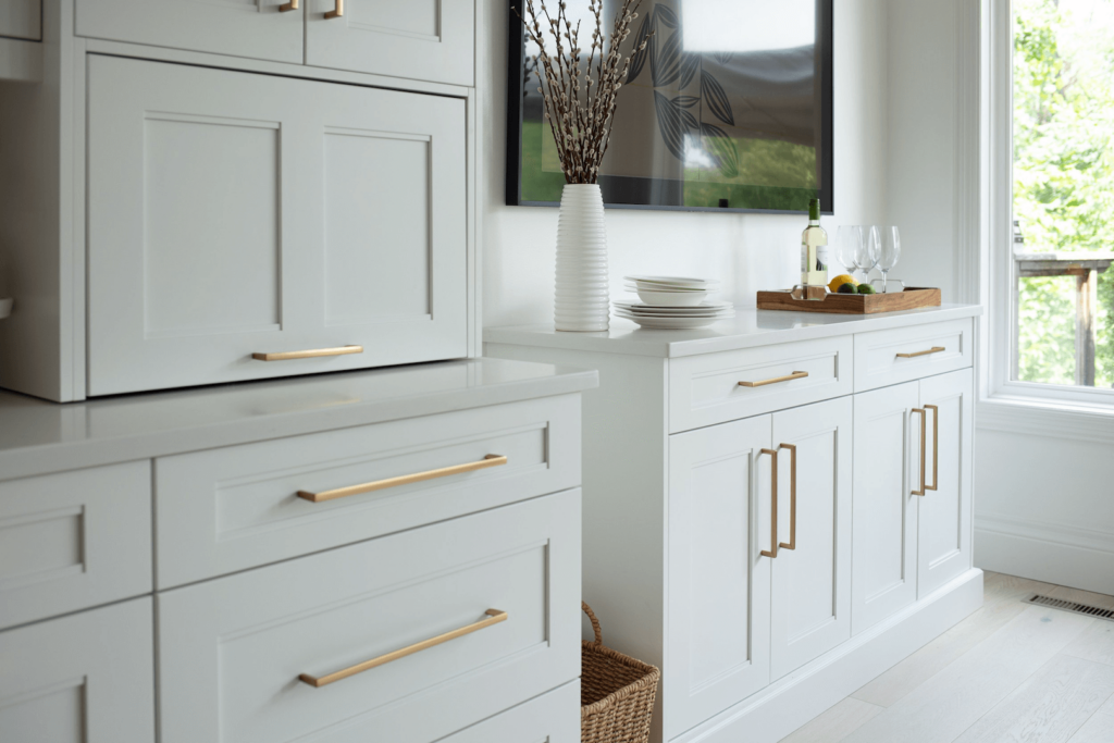 Z0ThX0fE White cabinetry with gold handles, a countertop with a wooden tray, and glassware arranged on a sideboard in a bright interior.